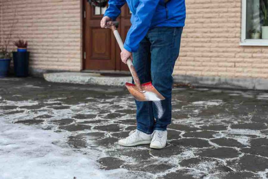 A man in blue jacket, jeans and white sneakers is using a shovel to sprinkle road salt