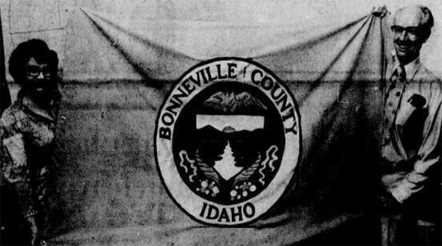 A. Wylie Starr, county commissioner, right, and Leola Mann, left, secretary, display Bonneville County's first official flag. The symbol shown, also found on the dome of the county building and county vehicles, is printed on a background of traditional orange. Caption dated July 3, 1977.