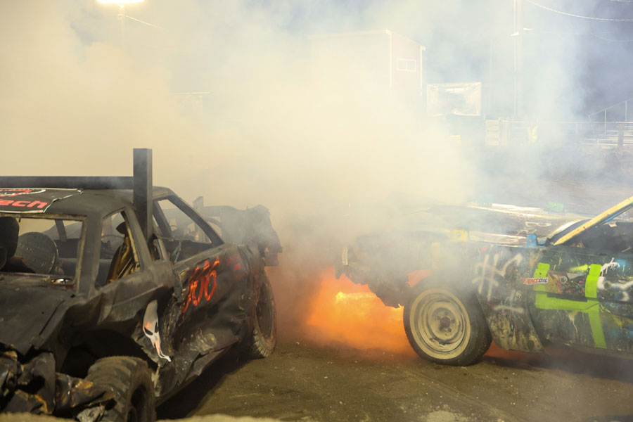 Drivers Cade Barney (666) and Devin Blood (47) battle as a nearby car catches fire in the Old Iron class at the 2025 Fall Brawl, held at the Fremont County Fairgrounds.