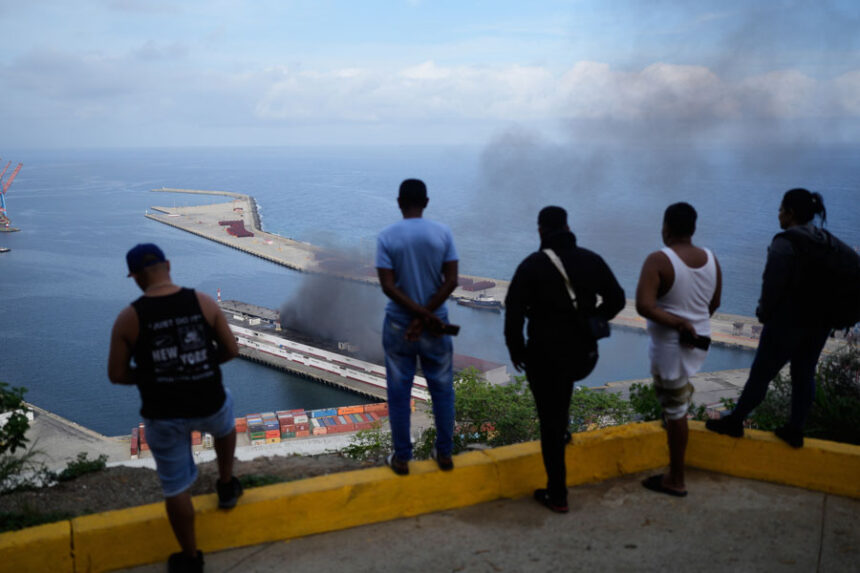 Men watch smoke rising from a dock after explosions were heard at La Guaira port, Venezuela, Saturday, Jan. 3, 2026. (AP Photo/Matias Delacroix)