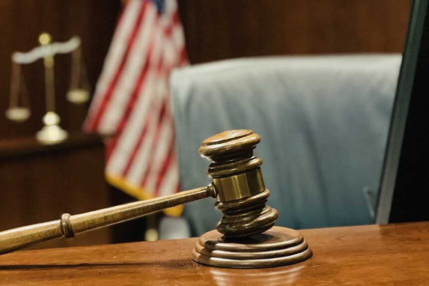 Selective focus on gavel, judge’s bench, scales of justice and flag in background.