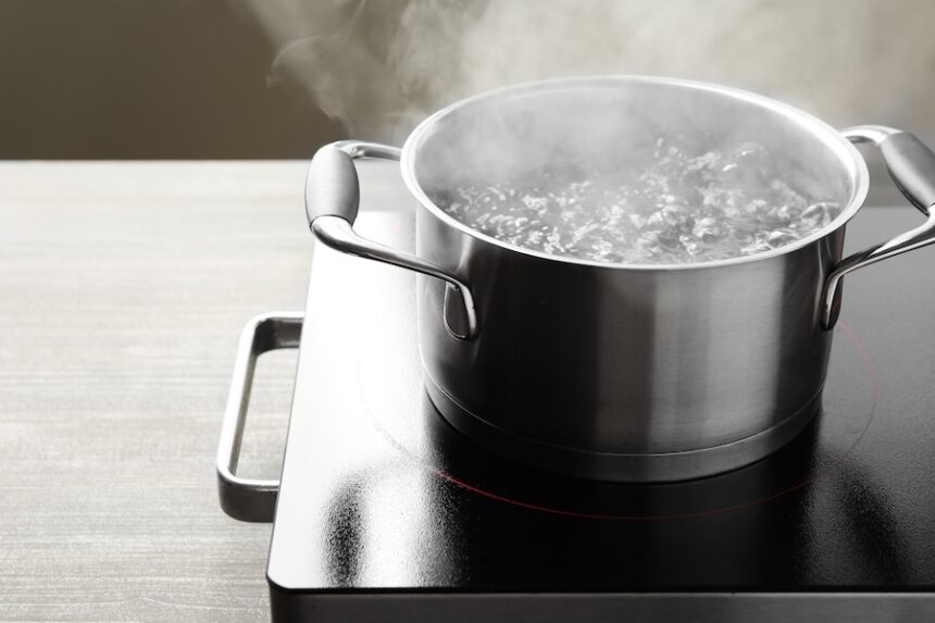 Cooking pot with boiling water and stove on wooden table against grey background, closeup