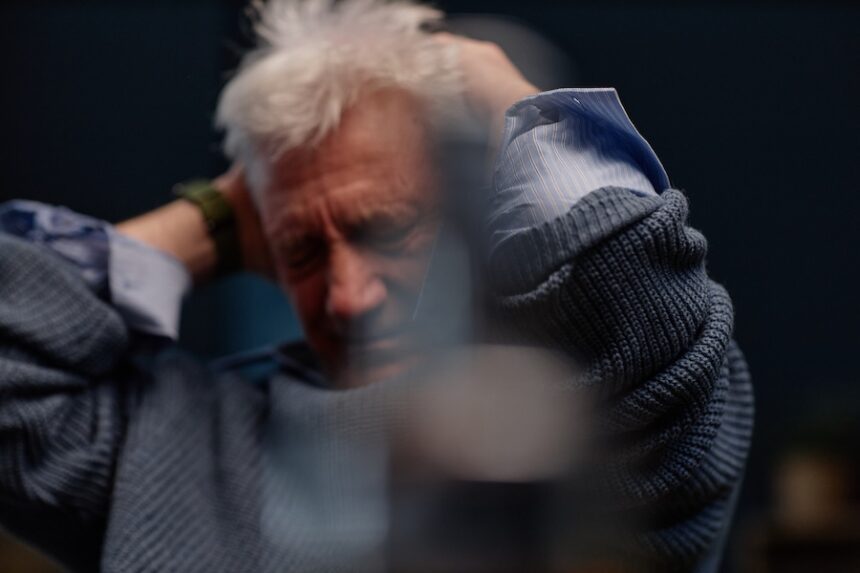 Elderly man with white hair holding his head in frustration, wearing a blue sweater without looking at camera. Visible indoors with abstract background and darkened lighting, dizzy spell