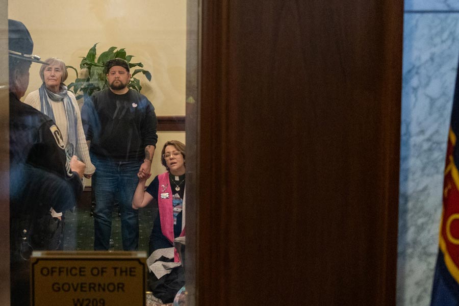 Trans advocate Nikson Mathews, center, listens to an Idaho State Police trooper moments before he and the Rev. Sara LaWall, right, were arrested in Idaho Gov. Brad Little’s office on Wednesday, April 1, 2026. | Photo by Kyle Pfannenstiel, Idaho Capital Sun
