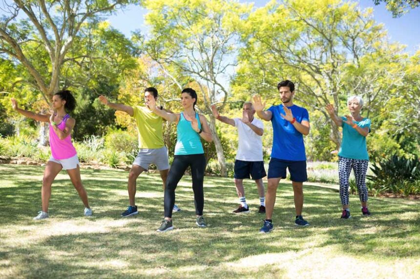 A group of seven people practice tai chi together in a verdant park on a bright, sunny day.
