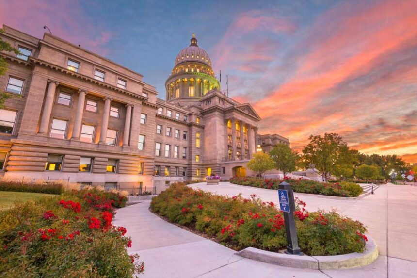 This photo captures a stately capitol building at sunset, framed by meticulously kept garden beds filled with red roses. The sky is filled with vibrant orange and pink clouds.