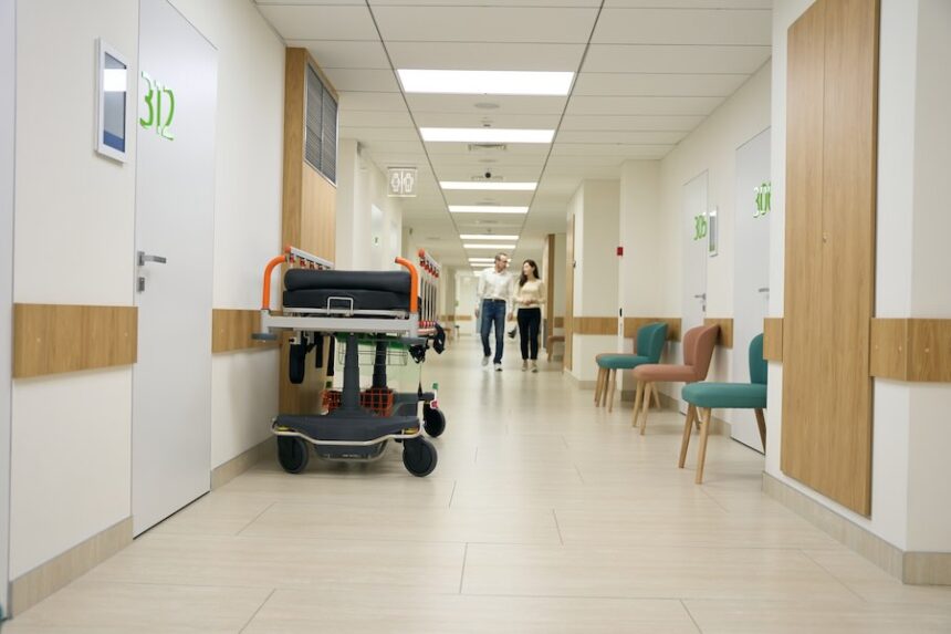Dark-haired woman and middle-aged man walk along the hospital corridor, in the foreground against the wall is a mobile bed. (Envato Elements)