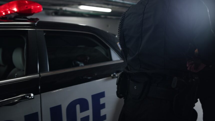 The photo shows a person dressed in a police officer uniform standing next to a police car inside a parking garage. The police car is black and white and has a red light on top.
