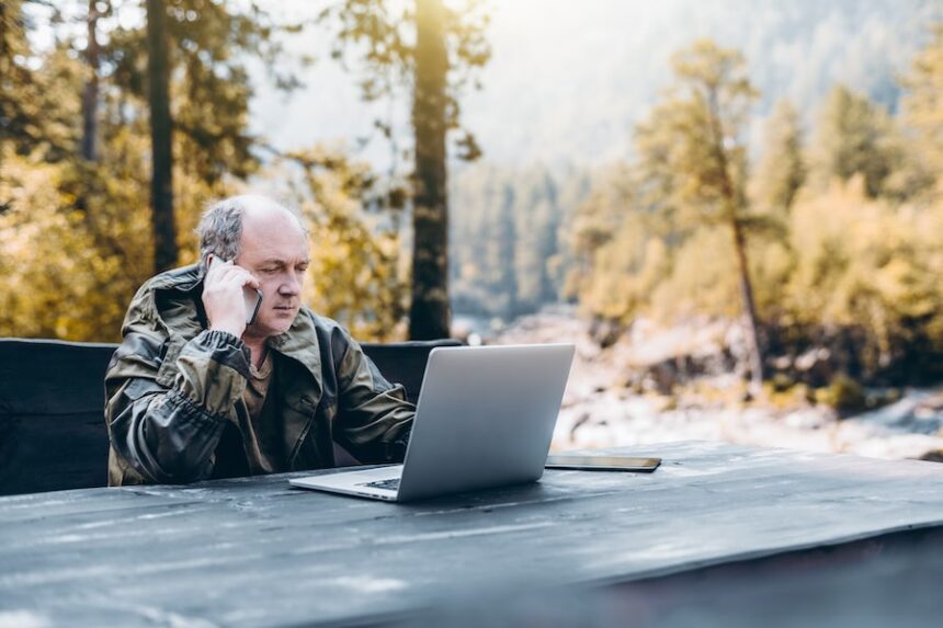 Serious pensive senior entrepreneur in work overalls is having a phone conversation while sitting at the wooden table with the laptop outdoors