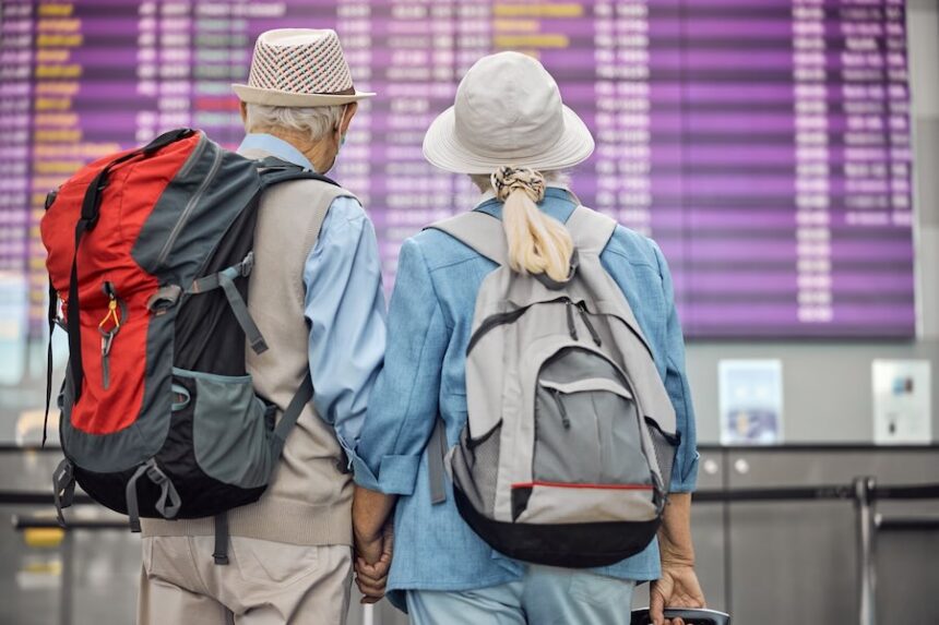 Back view of a senior couple with backpacks standing in front of the departures board.