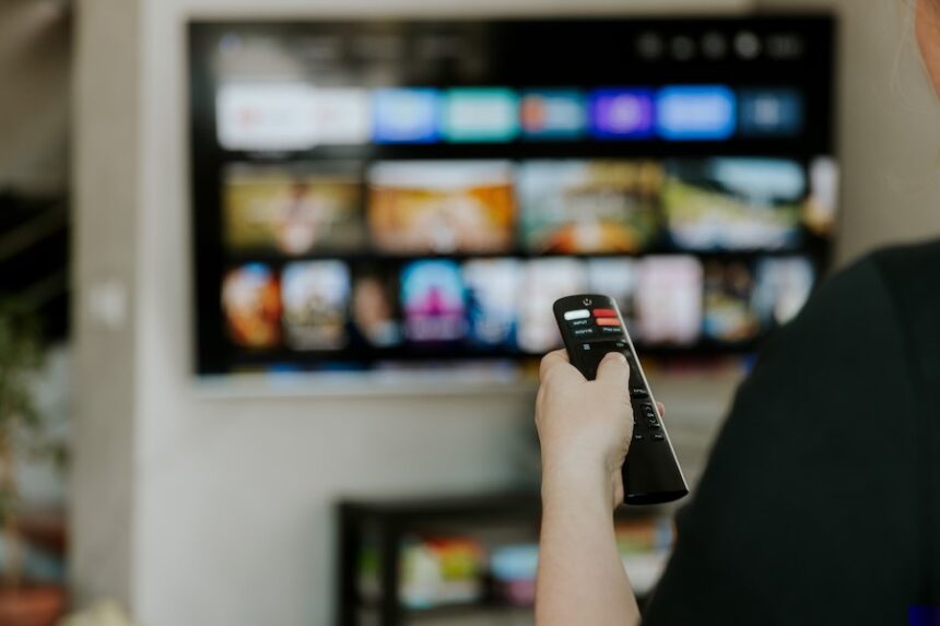 Hand holding remote control pointing to TV. Person watching smart television indoors. Blurred background. Colorful screen, copy space. Woman finger in foreground pressing button. Movie stream service. Netflix