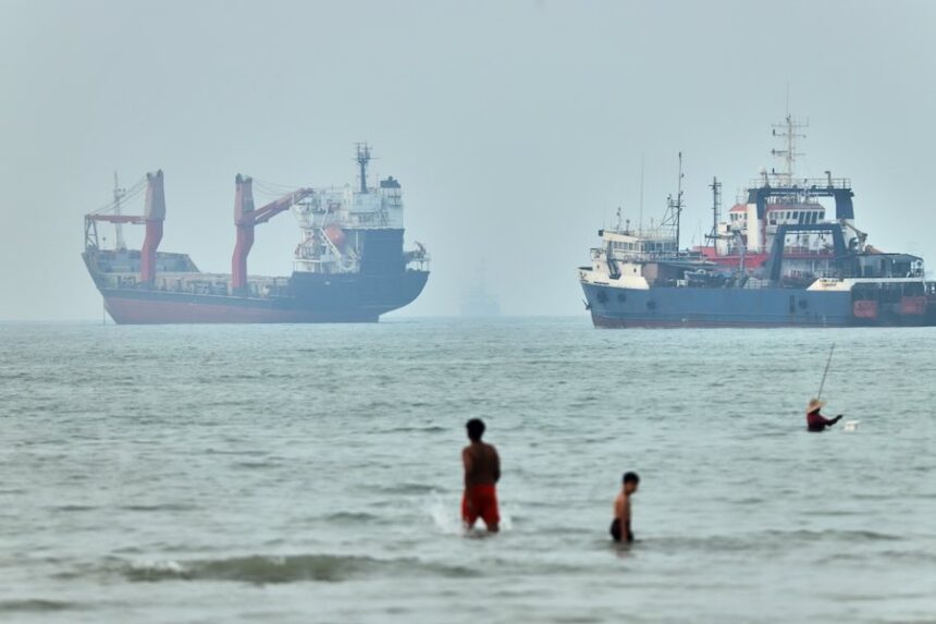 Children wade in the water with cargo ships at anchor in the background and a fisherman nearby, in the Strait of Hormuz off Bandar Abbas, Iran, Tuesday, June 30, 2026.
