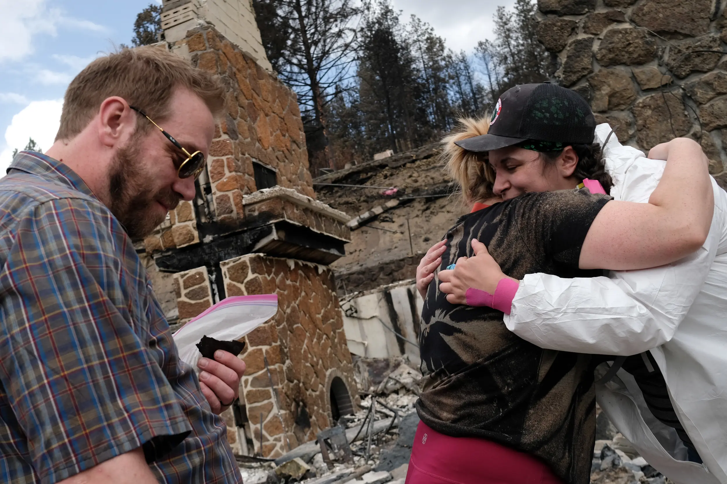 Keenan Burt, holds a bag containing the remains of his mother-in-law and a burnt drawing found nearby, as archeologist Kassie Rippee with Alta Heritage Foundation, right, embraces his wife CaitlLynn Burt, center on Saturday, Aug 29, 2026, after volunteer archeologists with Alta Heritage Foundation found some of the cremated remains of CaitlLynn Burt’s mother, which were lost the Old Trails fire Aug. 1, in Spokane, Wash. | Tyler Tjomsland, The Spokesman-Review