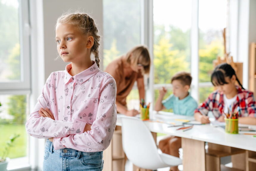 girl with braids standing with arms folded in classroom with students and teacher in background