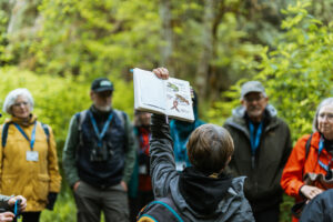 Woman holding up journal with nature drawings in front of group in forest.
