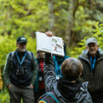 Woman holding up journal with nature drawings in front of group in forest.