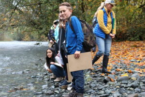 Group of kids standing on riverbank with woman in rainboots laughing beside them.