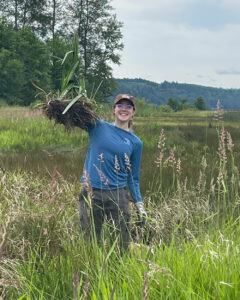 Young woman in grassy wetlands hoisting mass of dirt and plants