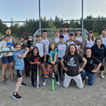 Group shot of adults and kids on a sandy baseball field.