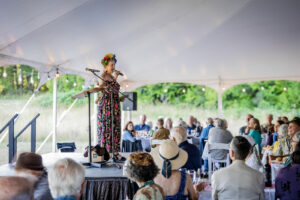 Woman in brightly colored dress on stage under large white tent addressing seated audience