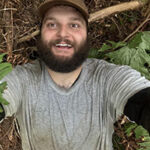 Selfie of young bearded man in baseball cap on forest floor