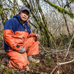 Japanese man in orange waders and rubber boots kneeling in forest clearing
