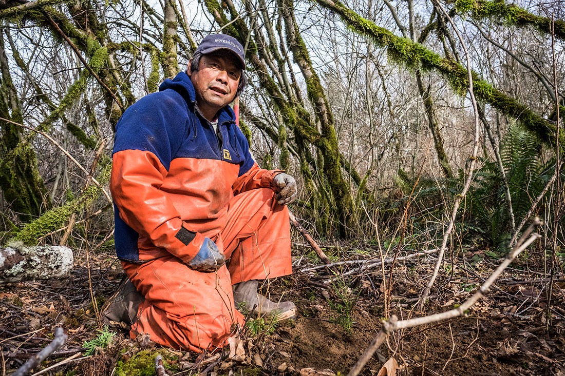 Japanese man in orange waders and rubber boots kneeling in forest clearing