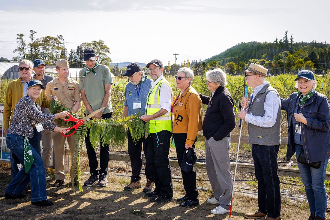 Group of people in front of flower field cutting cedar-bough "ribbon" with huge scissors.