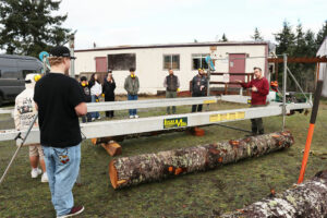 Students standing around sawmill and large log outside near school portable building.