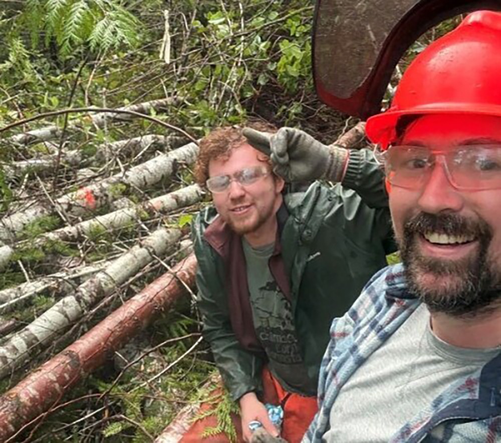 Photo of two men with a pile of logs behind them.