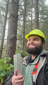 Photo of a man with a yellow hard hat holding a clipboard in a forest