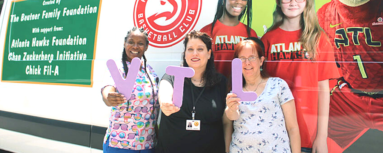 three women holding large letters spelling "VTL" stand together in front of the Vision to Learn van
