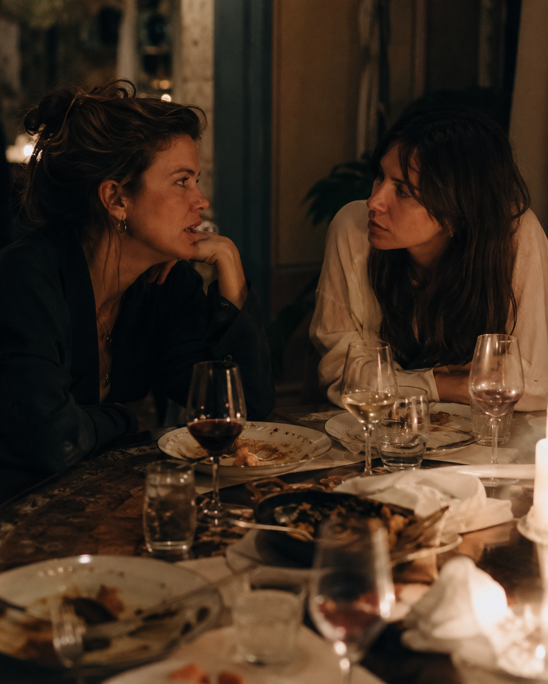 Two women in close conversation across a table late in the evening after a shared meal
