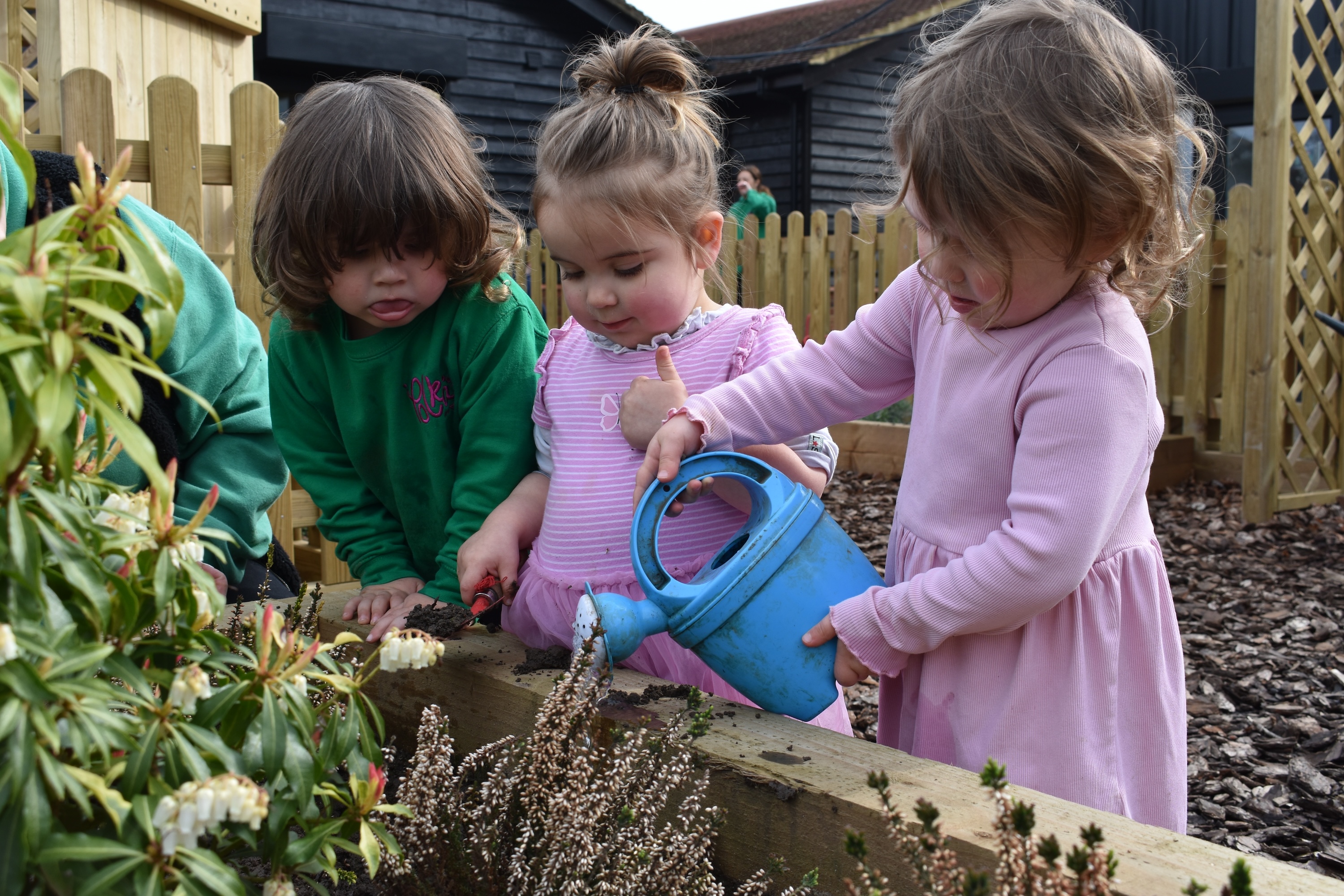 Three early years children learning through play in an EYFS educational setting, watering plants and shovelling soil too.