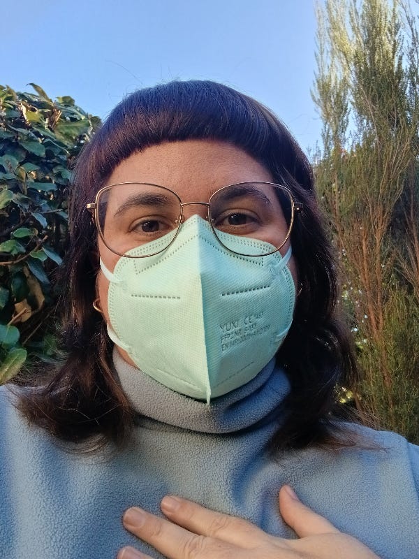 Photo de moi devant un ciel bleu et des buissons. Je porte un masque FFP2 bleu lagon et un col roulé bleu ciel.