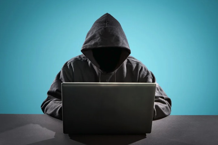 Person in black hoodie sitting behind a computer with face shadowed