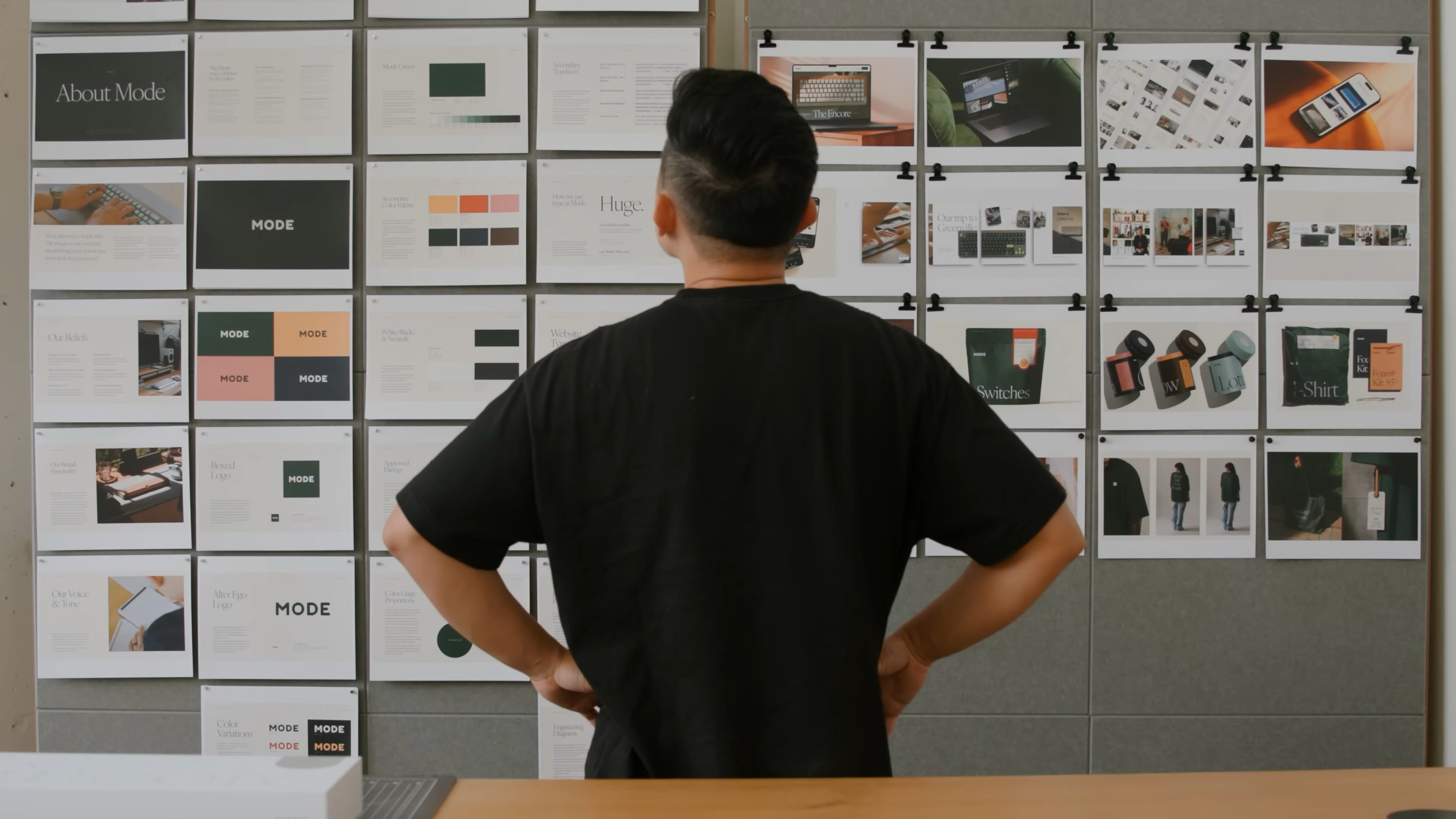 A designer stands in front of a large wall covered in Mode brand identity mockups, reviewing logo designs, color palettes, packaging, apparel, and digital touchpoints. This scene visually captures the holistic approach to branding and the decision-making process featured in the “Building a Brand – Redesigning a Business Start to Finish” video, highlighting how every element works together to create a cohesive visual system.