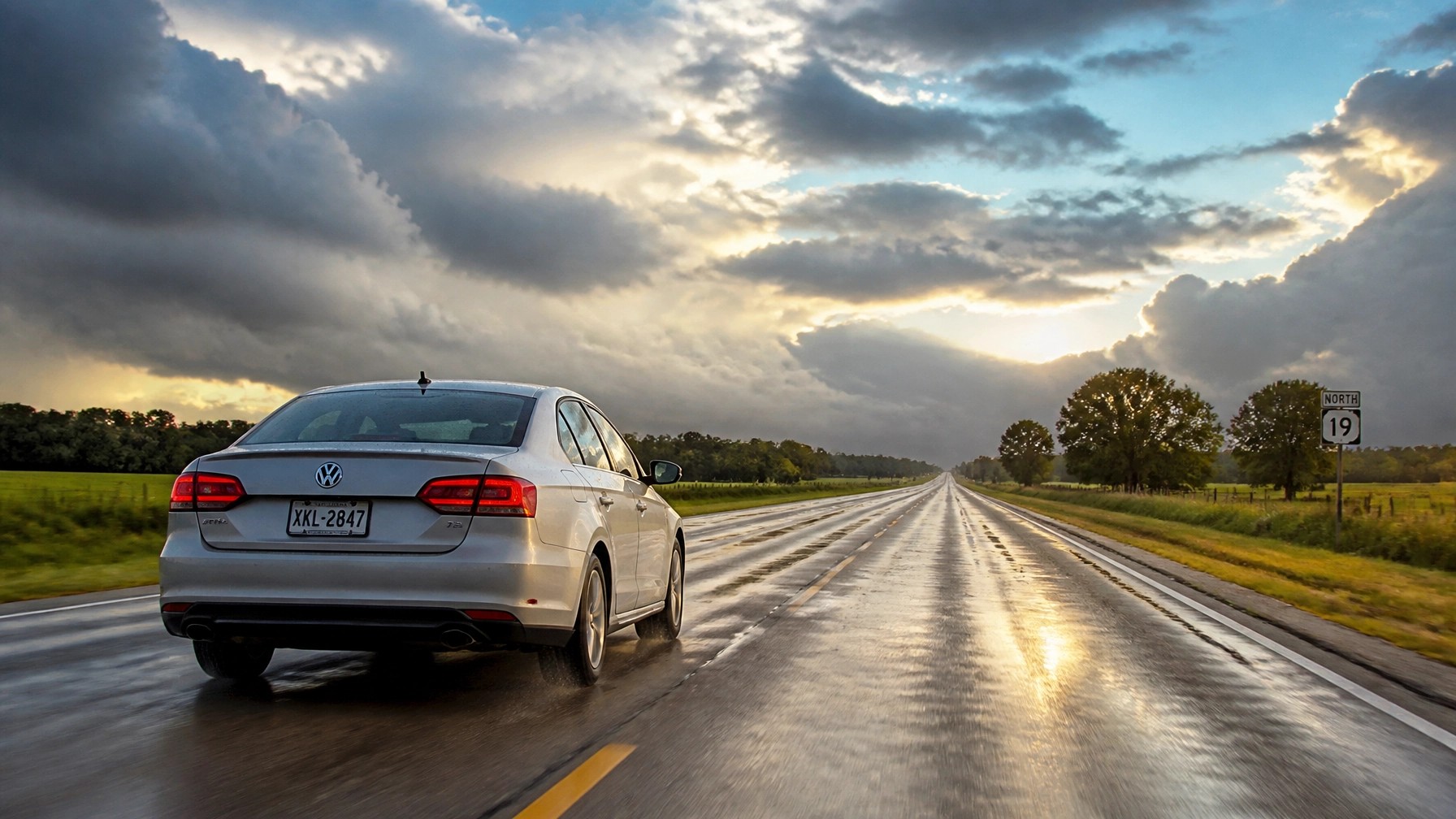 A silver Volkswagen Jetta driving on a long, wet highway in the rural American South under a dramatic post-storm sky.