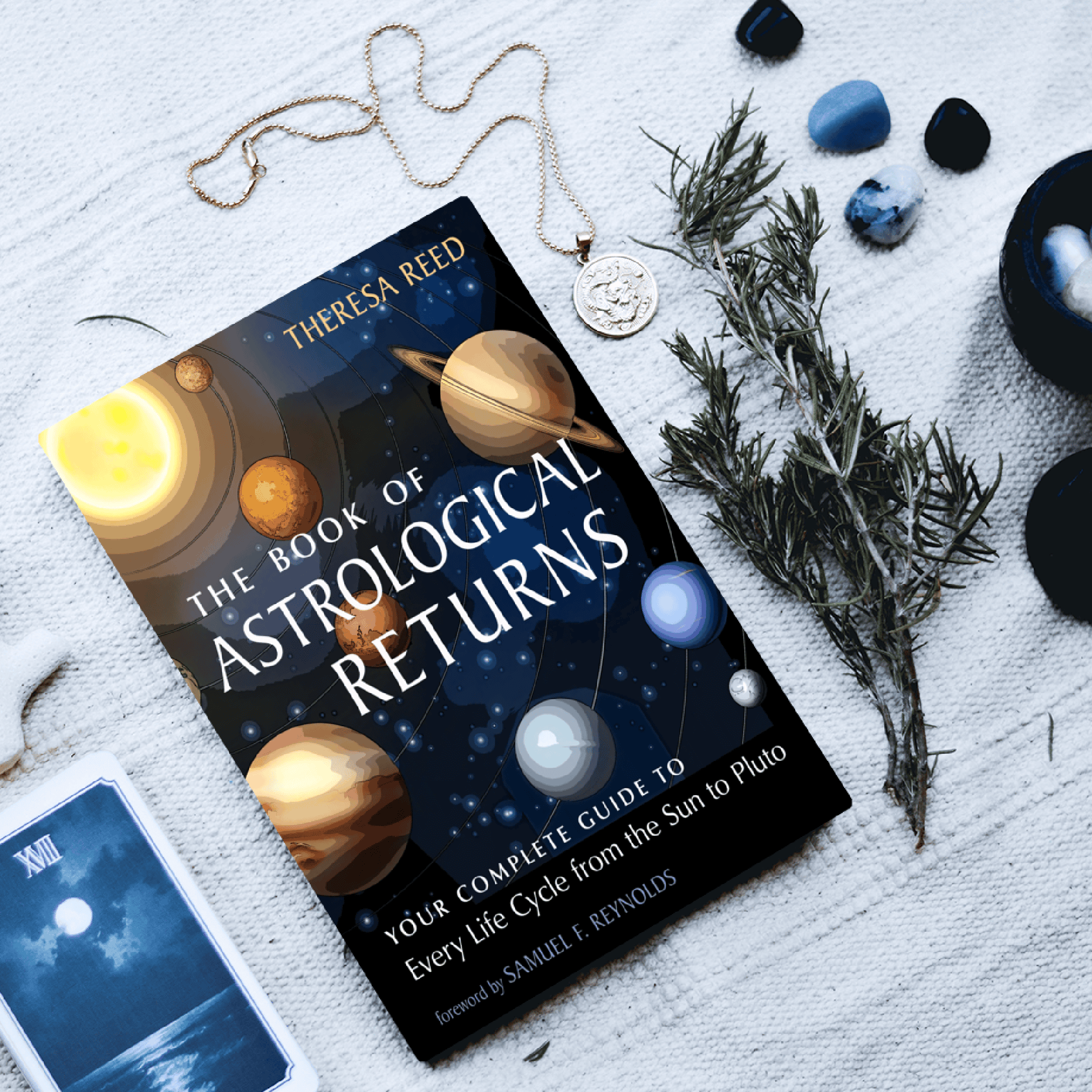 The Book of Astrological Returns by Theresa Reed on a table surrounded by a Tarot card, an amulet, and sprig of pine