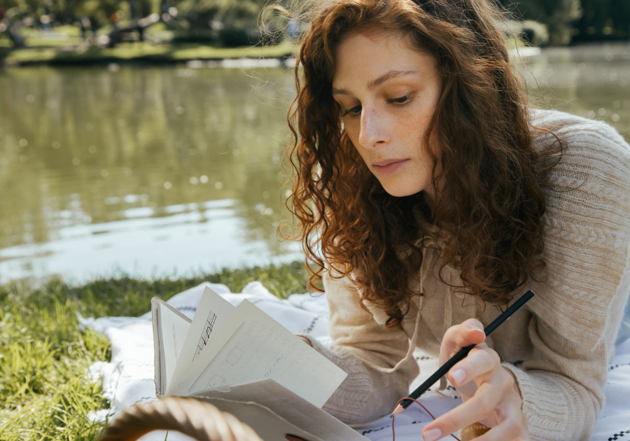 A young lady laying on a blanket in the grass outdoors journaling.