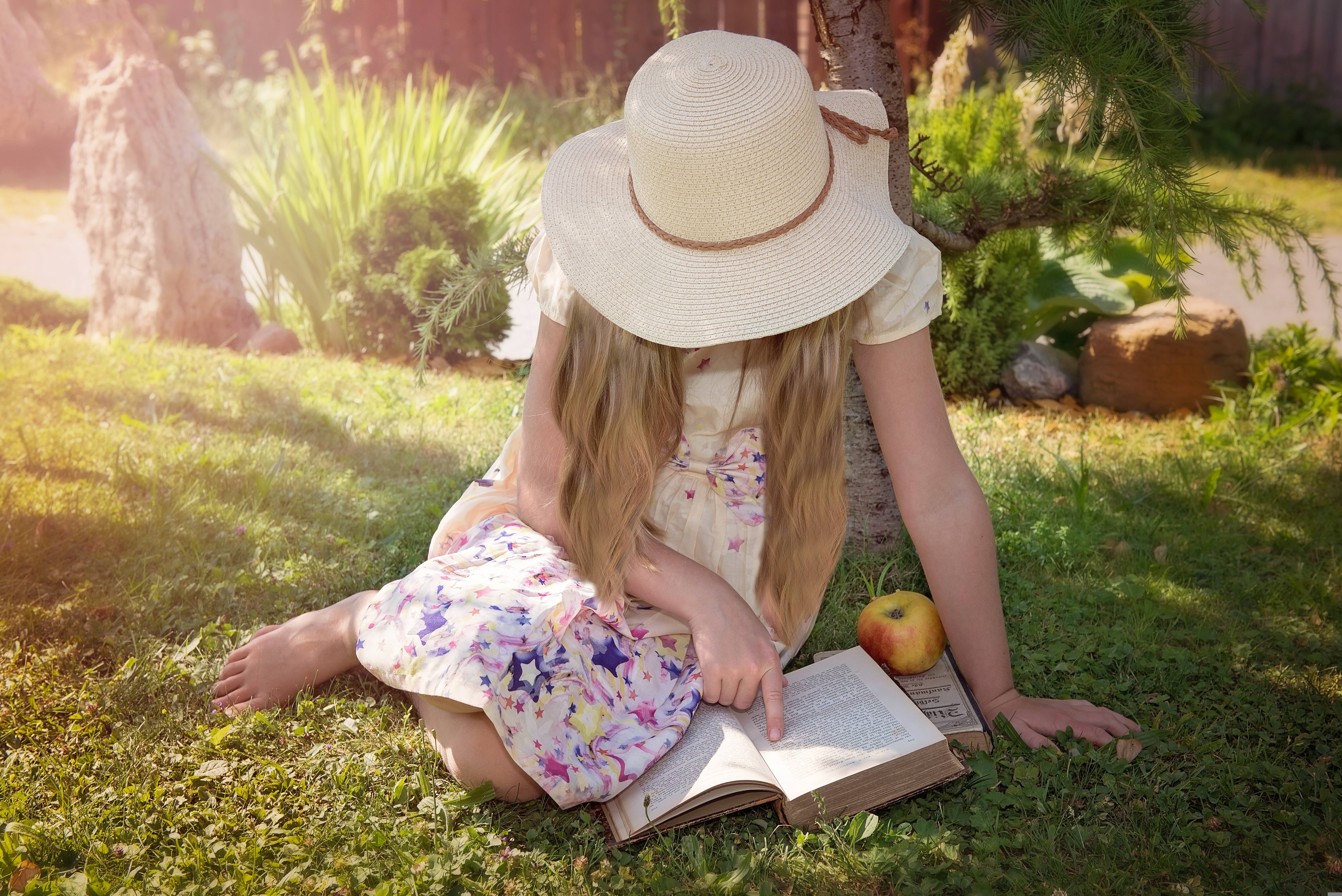 little girl with a large-brimmed hat on, sitting on the grass under a tree, reading a book