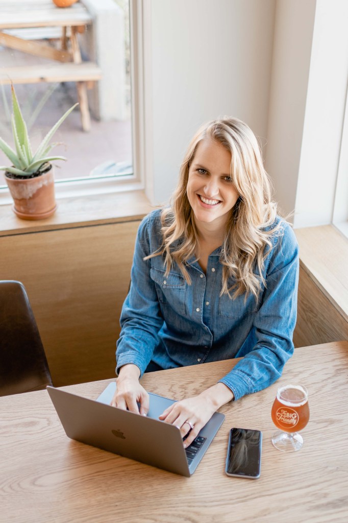 Kayla, sitting at a minimalist desk with a beer, her computer and phone, this is how moms work