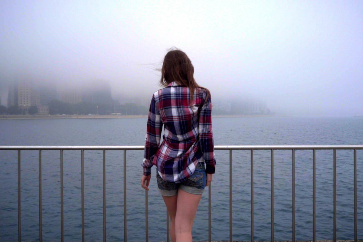 A woman pictured from behind staring out in to a body of water, fog above. 