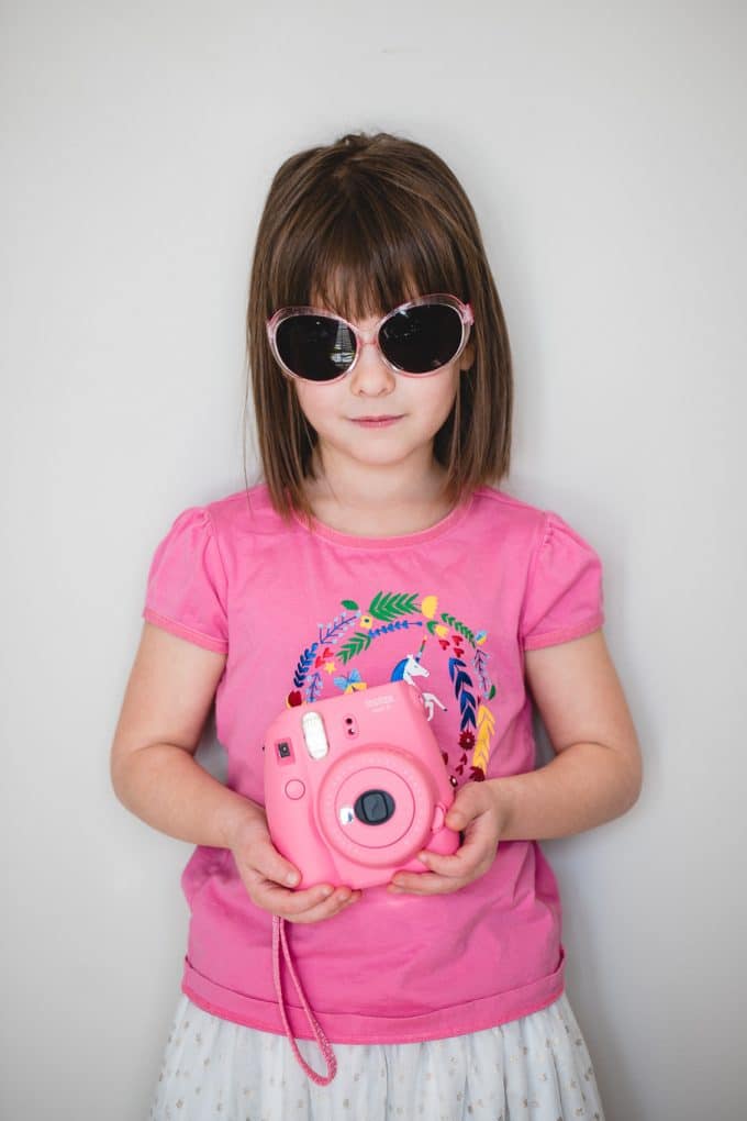 Child wearing pink sunglasses, pink shirt, and holding a pink camera in front of a white wall. Photography for kids is easier than you might think.