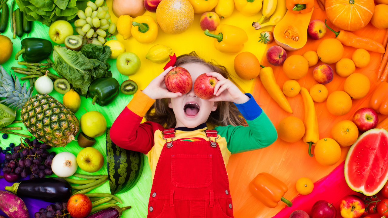 A happy little girl, holding red apples over her eyes, laying down amidst all sorts of fresh fruits and vegetables.