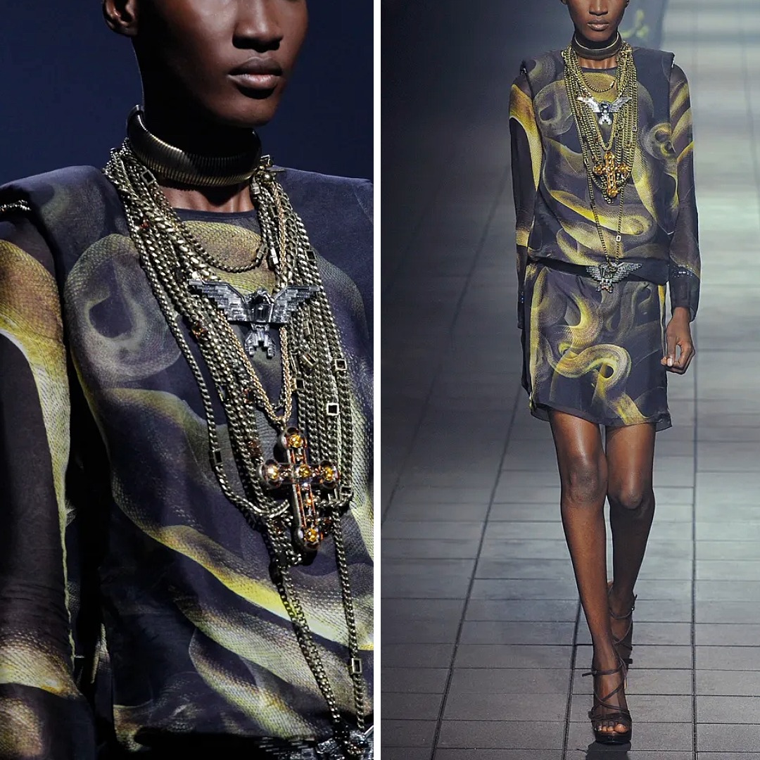 Runway look from Lanvin Fall 2012 featuring a sheer black dress with a yellow smoke-inspired print, styled with multiple layered metal chains and cross pendants. The outfit shows a softer interpretation of black-and-yellow contrast through print and accessories.