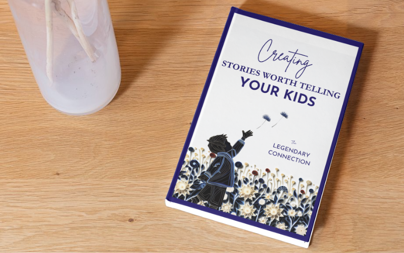 Flat lay of the book "Creating Stories Worth Telling Your Kids" by The Legendary Connection resting on a wooden surface next to a minimalist vase, with a cover illustration of a child blowing dandelion seeds among whimsical paper-quilled flowers.