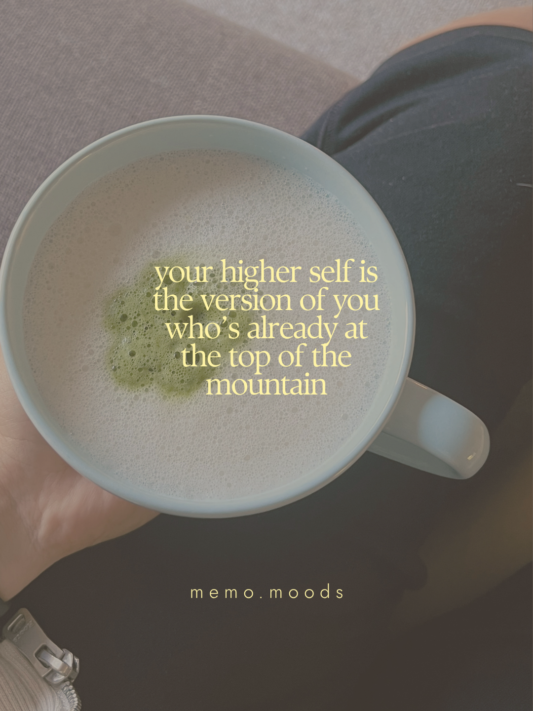 Quote graphic over an overhead shot of a matcha latte in a white ceramic mug held by a person sitting on a couch. Text reads: "your higher self is the version of you who's already at the top of the mountain." by memo.moods.