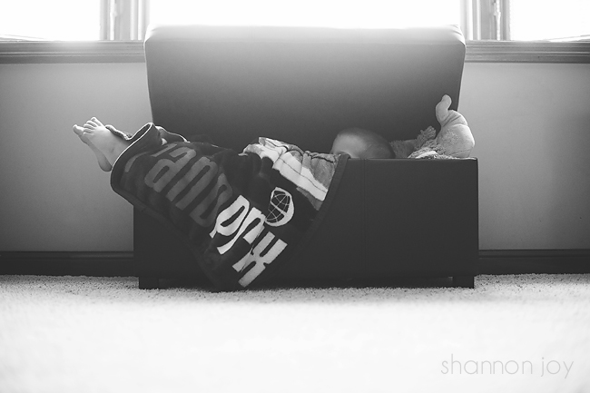 Black and white photo of a child laying in a toy box in front of a bright window during project 365