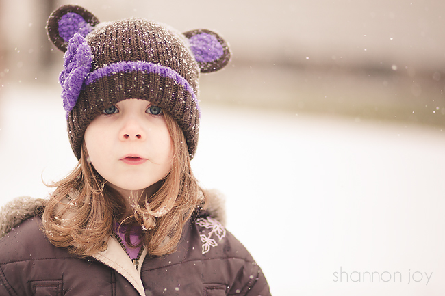 Photograph of a child in the snow wearing a brown hat with purple ears